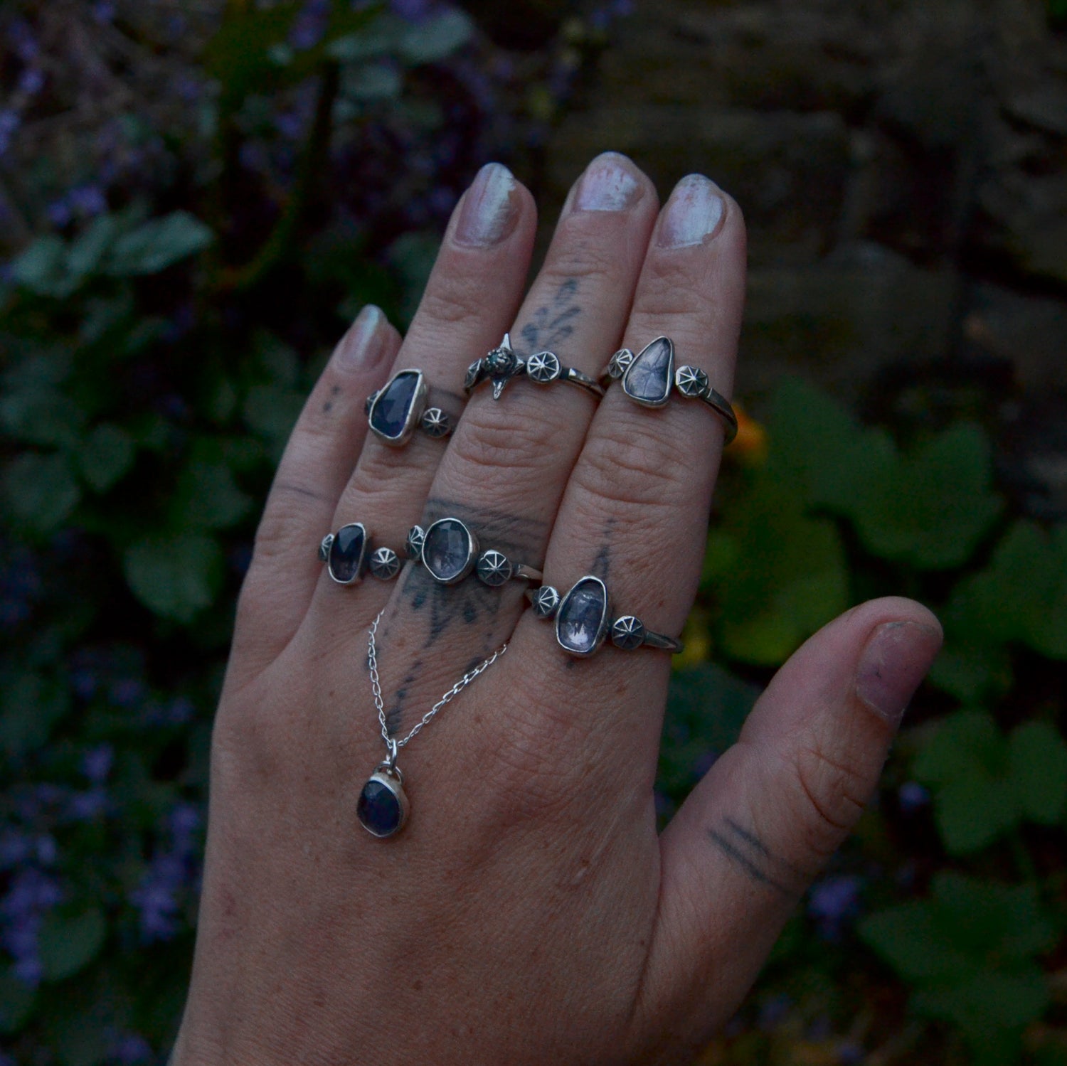 Close-up of a hand adorned with five silver rings and a silver necklace featuring blue and translucent gemstones. The rings and necklace have unique, handcrafted designs, with intricate details and floral motifs. The hand has visible tattoos on the fingers, and the nails are painted with a light metallic polish. The background is slightly blurred with green foliage and flowers, highlighting the jewellery as the focal point.