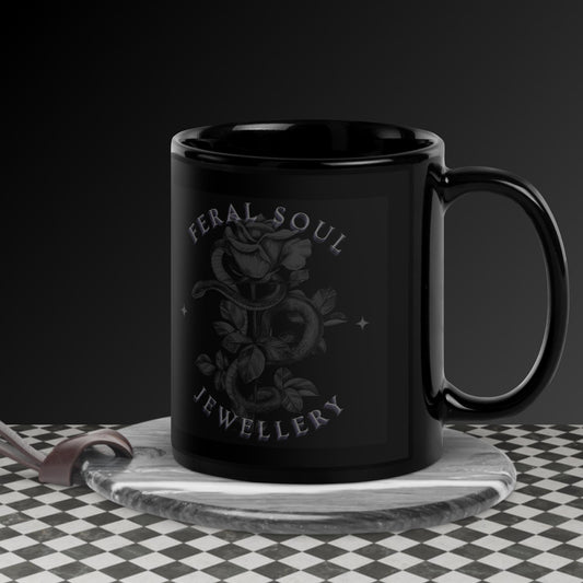 Black ceramic mug featuring a gothic design with a rose and snake illustration. The text "FERAL SOUL JEWELLERY" is printed in a curved, stylised font above and below the image. The mug is placed on a dark surface with a chessboard in the background.